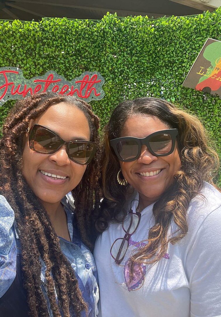 Two momz in sunglasses pose for the camera in front of a Juneteenth hedge.